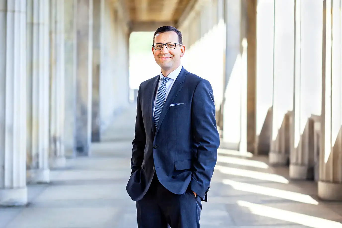 Porträt eines Mannes in einem eleganten dunkelblauen Anzug mit Krawatte, der lächelnd in einem Säulengang steht. Das natürliche Licht und die architektonischen Details im Hintergrund schaffen eine professionelle und stilvolle Atmosphäre. Aufgenommen von Concept Photography Berlin, vermittelt das Bild einen Eindruck von Kompetenz und Souveränität.