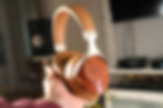 Hand holding brown headphones with wooden accents marked "SIVGA", set against a blurred indoor background with audio equipment.