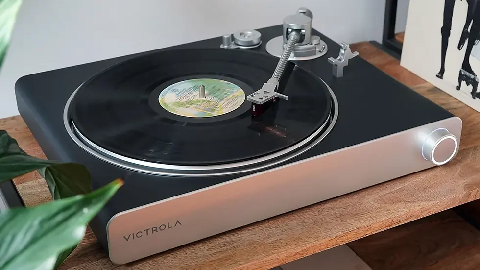 Turntable playing a record, with "Victrola" brand visible. Set on a wooden table, surrounded by plants and vinyl albums. Calm, vintage vibe.