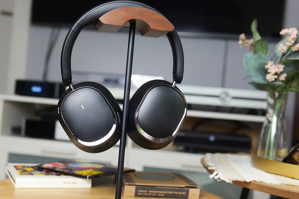 Black headphones on a wooden stand with albums and books nearby. Background shows a TV, console, and a vase with flowers.