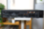 Back panel of a black audio device with various ports and connectors, including HDMI and RCA, set on a white stand against a blurred room.