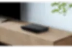 Black Sony Blu-ray player on a wooden table, next to a TV. Green plant in background. Minimalist and modern setting.