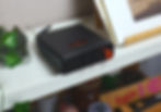 Black audio amplifier with an orange knob on a white shelf. Surrounded by a green plant in a glass vase and a framed picture. Shelving holds various items with Asian designs.