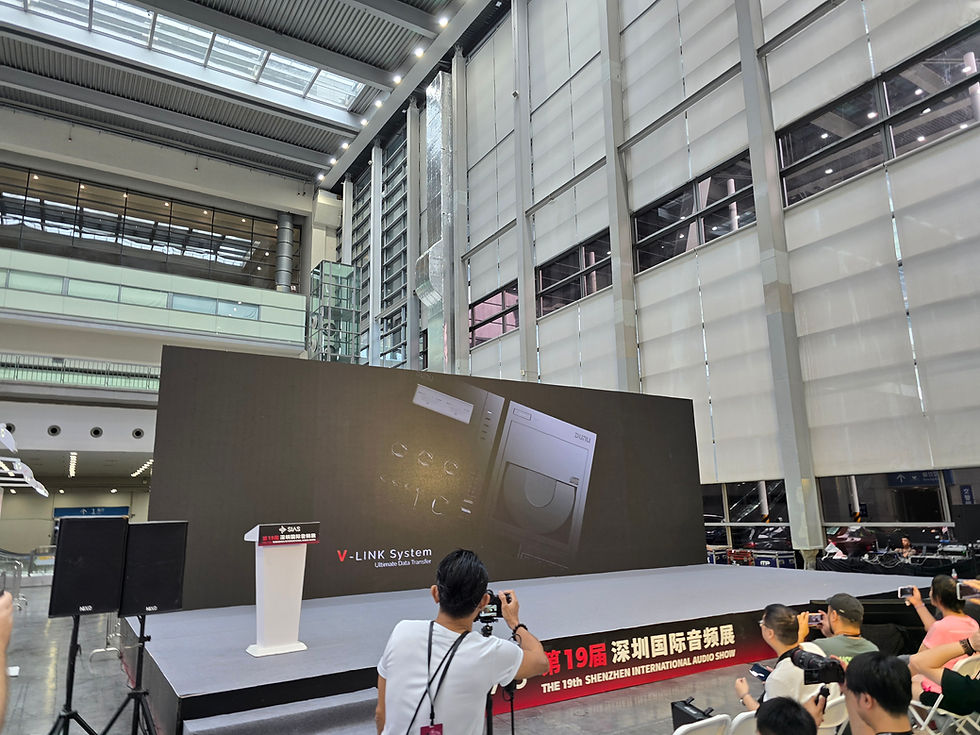 Stage with podium and large screen displaying "V-LINK System" at a tech event. Audience seated in modern, spacious venue.