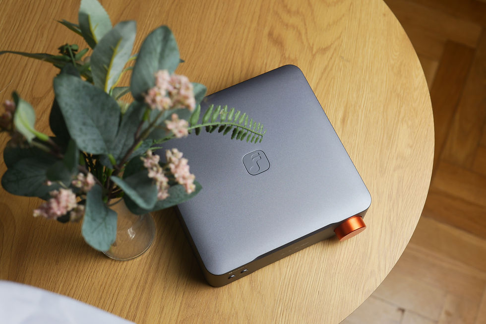 Gray compact device with orange knob on a wooden table, partly covered by a vase of green leaves and pink flowers.