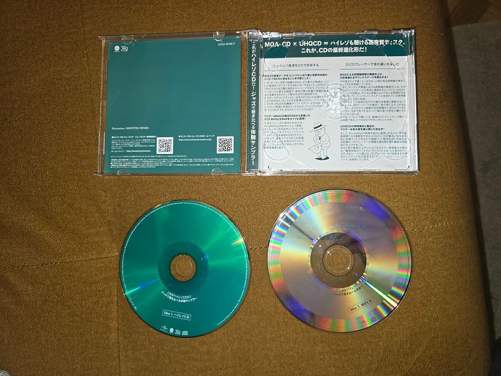 Open CD case with Japanese text and two discs on brown fabric: teal hybrid CD and reflective silver CD with rainbow sheen