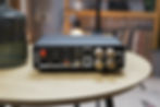 Black audio amplifier on a wooden table, showing various input and output ports. Text: "MADE IN CHINA". Blurred room background.