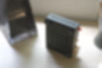Black electronic device on a light wood surface, featuring multiple connectors and a red knob. Nearby are shadows and a metallic object.