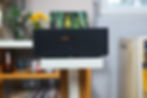 Black Marantz speaker on a white stand, background shows bookshelves and flowers in vases. Soft, natural light fills the room.