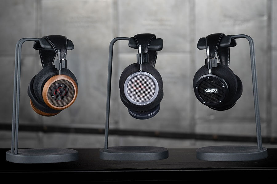 Three headphones on stands in a studio setting: wooden, silver, and black designs. "GRADO" visible on the right headphone. Gray backdrop.