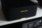 Black Marantz speaker on a shelf with glowing logo. Minimal controls visible. Sleek design in a dimly lit room. Silent ambiance.