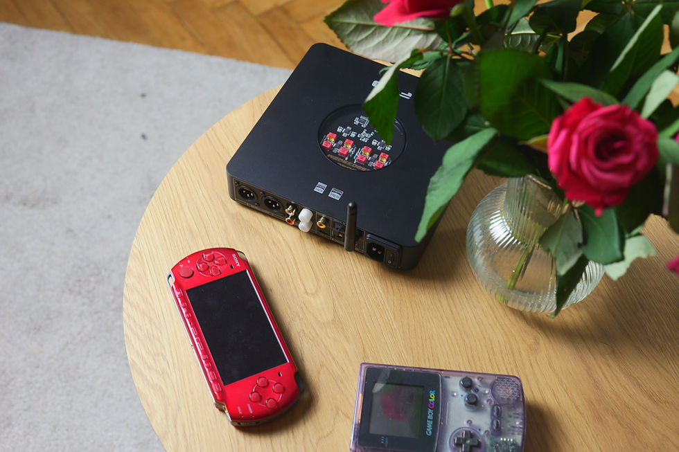 Red PSP and translucent Game Boy Color beside a black audio device on a wooden table, with pink roses in a vase.