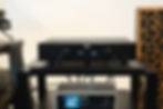 A black audio amplifier displays "XLR 24" on a shelf, with a modern stereo below showing "Fade To Black" by Dire Straits. Beige panel backdrop.