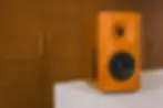 Orange speaker on white marble surface against a wooden cabinet background. Speaker design is sleek and modern. No text present.