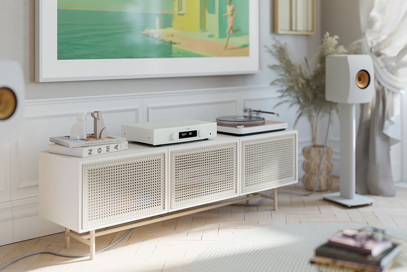 Minimalist living room with a white sideboard, a record player, and speaker. Art print on wall, potted plant, and natural light enhance serenity.