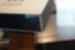 Close-up of a silver music streamer on a wooden table. Features a black front panel with two input jacks. Text reads "MUSIC STREAMER."