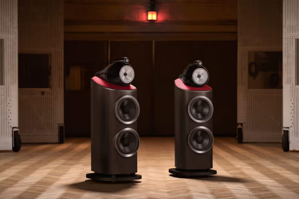 Two sleek speakers on a wooden floor, featuring a black finish with red accents. They stand in a warmly lit room, no text visible.