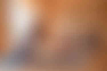 man teaching yoga to two women in a wooden studio, with straps on the wall and a large window. All are seated in a forward reaching stretch pose on blue yoga mats.