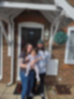 Family of three smiling in front of a brick house with a dark door. The baby is in floral pajamas. Hanging plant and light visible.