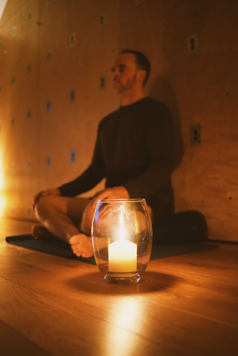 A man sits meditating in a dimly lit room with wooden walls, a glowing candle in a glass holder visible in the foreground.