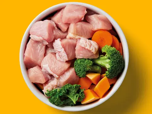 Bowl of chicken, broccoli, and carrots on yellow background, for dog food