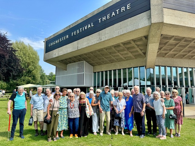 Group of smiling elderly people outside Chichester Festival Theatre on a sunny day. Building with glass windows and trees in the background.