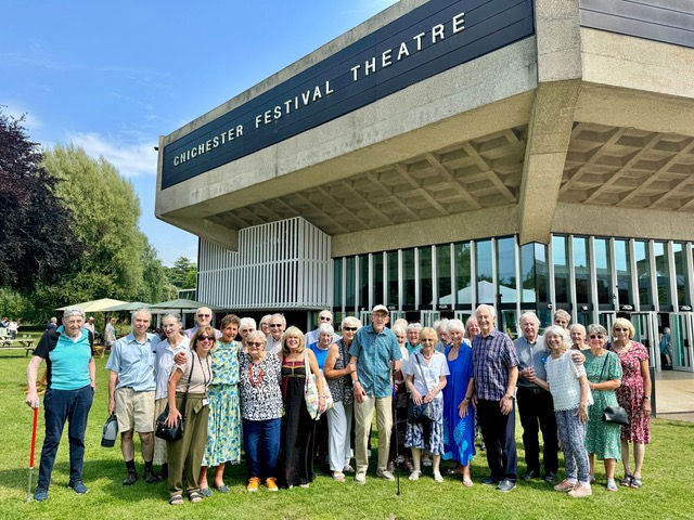 Group of smiling elderly people outside Chichester Festival Theatre on a sunny day. Building with glass windows and trees in the background.