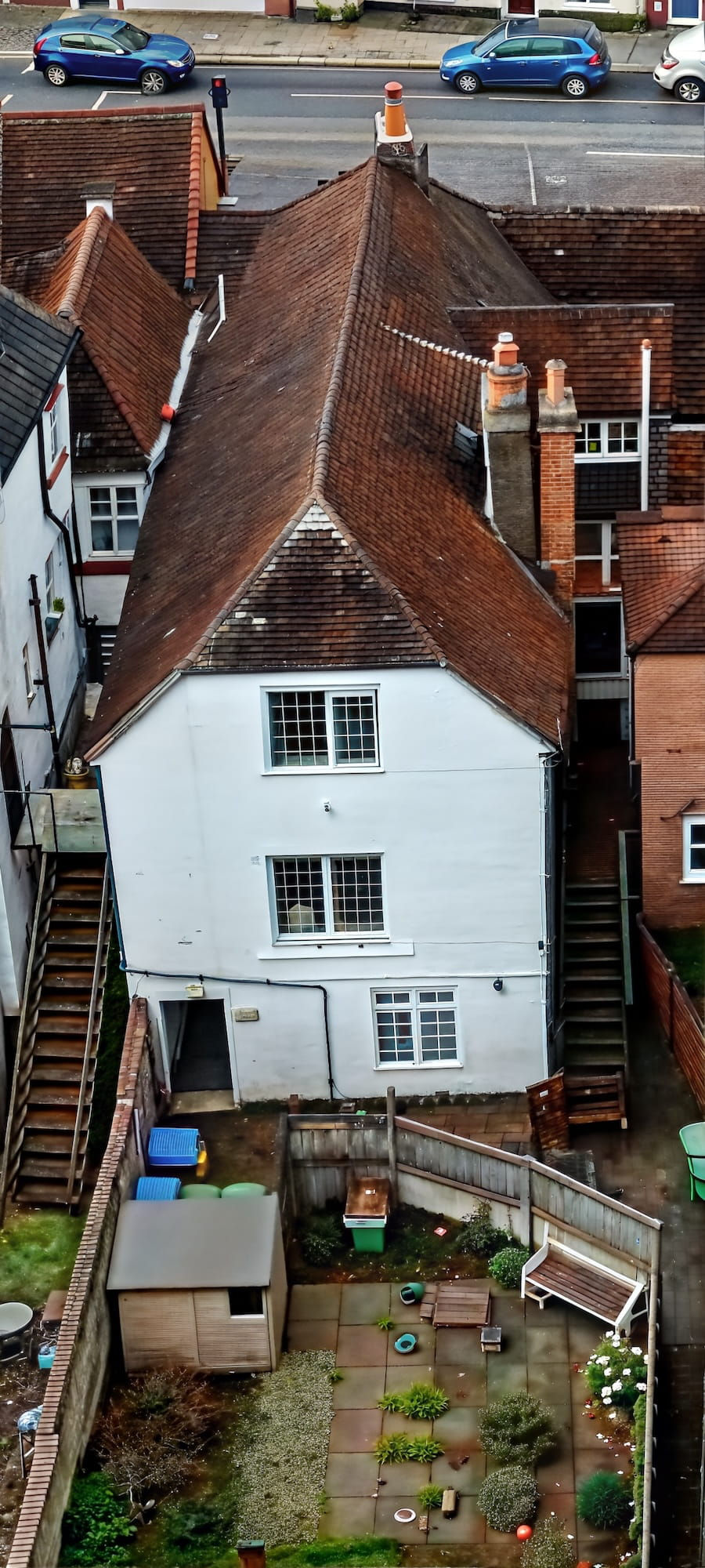 Aerial view of a white house with brown tiled roof. Backyard has a shed, benches, and colorful bins. Street with blue cars in the background.