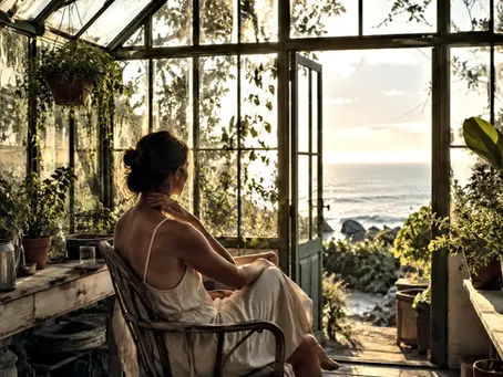 Woman seated inside a sunlit coastal greenhouse overlooking the ocean, surrounded by plants and warm summer light, reflecting quietly at golden hour.