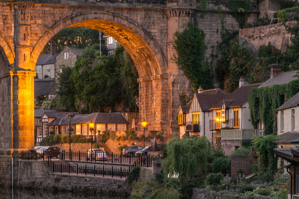 Stone arch Knaresborough Viaduct lit by warm lights in the evening. Below are parked cars and quaint Yorkshire houses, surrounded by lush greenery. Peaceful mood.