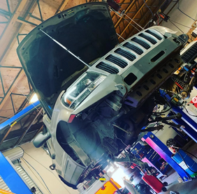 Grey Jeep lifted for auto repair with open hood in garage.