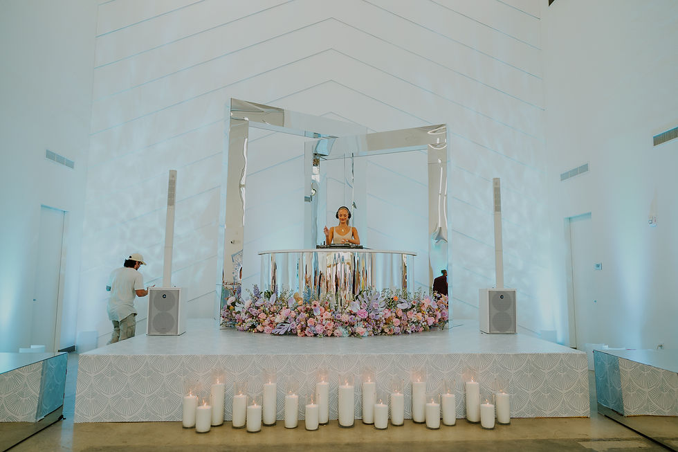 DJ in white setting plays music behind silver booth, surrounded by pastel flowers and candles. Person adjusts speaker. Calm atmosphere.