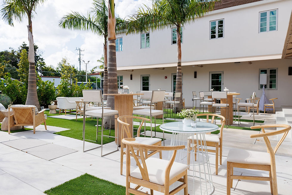 Outdoor patio with wooden tables, chairs, palm trees, and a white building in the background. Sunny, inviting atmosphere.