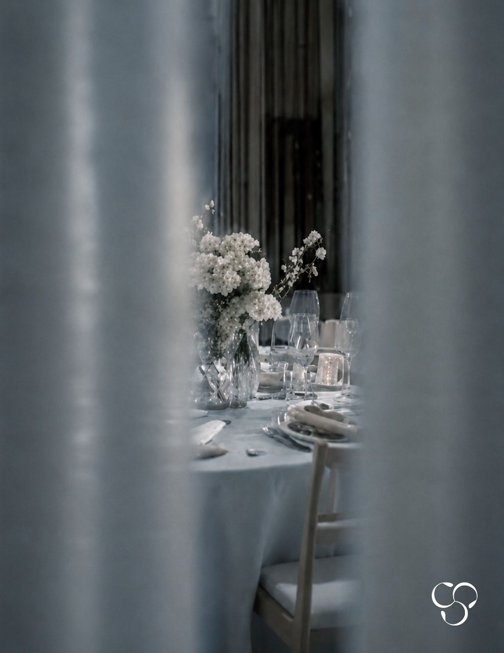 Elegant reception table seen through sheer curtains, with white floral centerpiece, glassware, candles, and soft gray lighting.