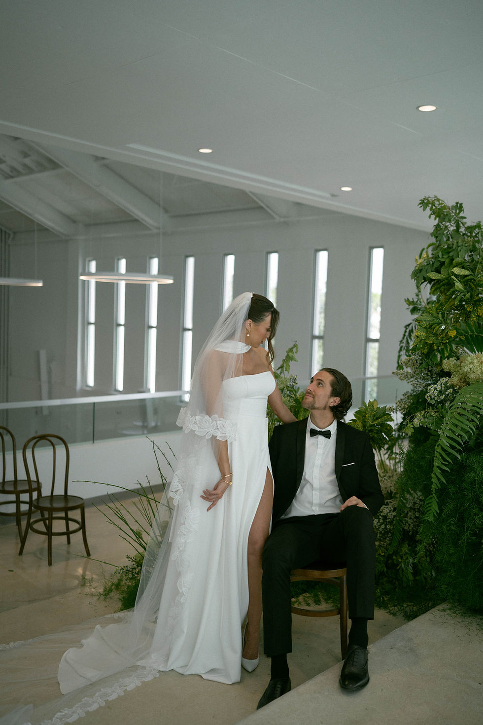 Bride in a white gown stands beside a seated groom in a black suit. Indoor setting with plants and modern lighting. Romantic mood.