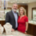 Smiling man and woman in a jewelry store, standing near a display of necklaces and bracelets. The woman wears a red dress.