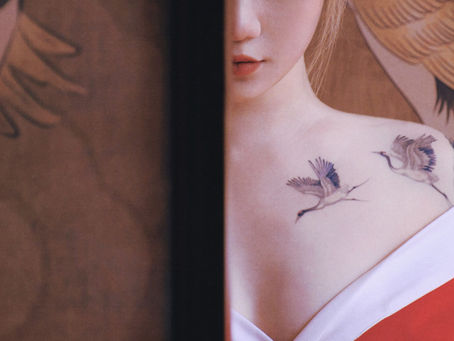 A delicate fine-line crane tattoo on the collarbone and shoulder, featuring two red-crowned cranes in flight rendered in soft black ink, photographed against the warm tones of a traditional East Asian painting. Tattoo by Wild Crane Tattoos, Markham ON.