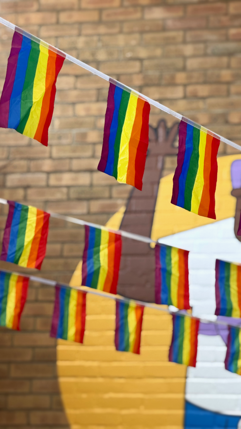 A photo of rainbow flag bunting in front of a bare wall with a mural. The bunting overlaps three times and the line at the front is in focus.