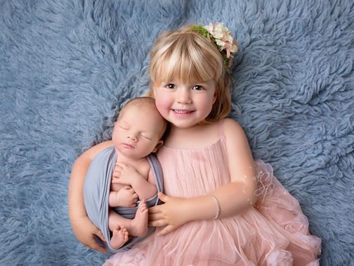 A heartwarming portrait of a young girl gently holding her newborn sibling, both dressed in soft pastel tones, capturing the tender bond between siblings.