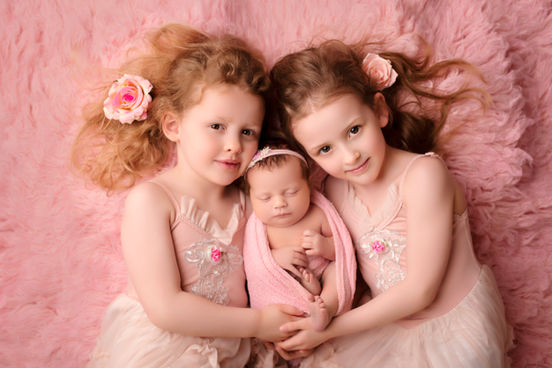  A loving portrait of two sisters embracing their newborn sibling, all dressed in soft pink gowns, highlighting their bond and the warmth of family.