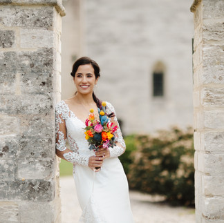 A bride in a lace wedding gown holding a colorful bouquet, framed by a historic stone archway at Stevns Klint.