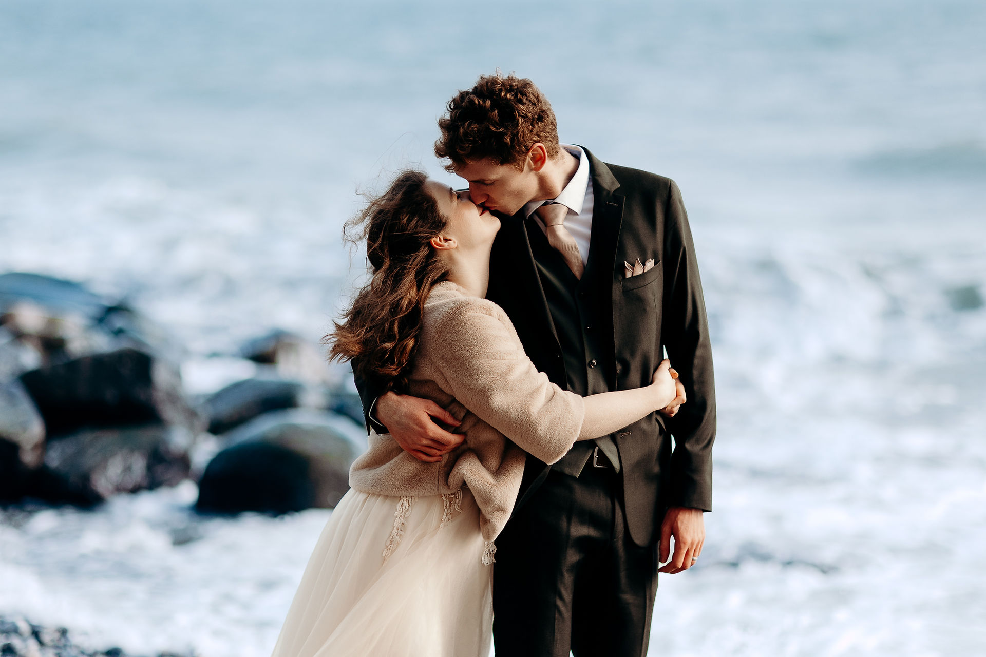 A romantic kiss between a bride and groom on a windy day by the sea, with waves crashing against the rocks.