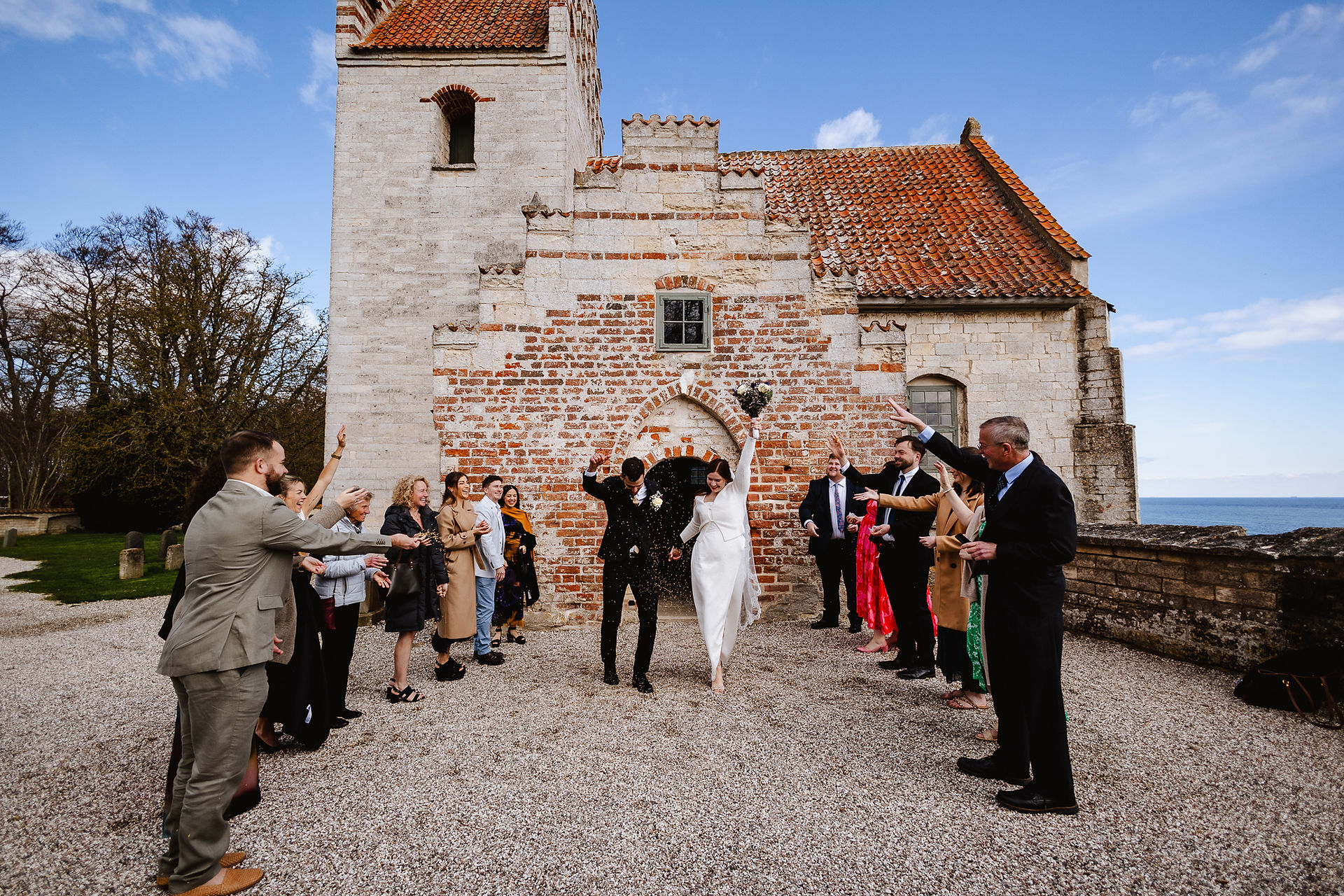 A joyful wedding couple walking out of the historic Højerup Old Church while guests celebrate with flower petals.