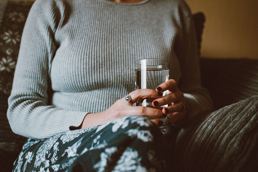 Woman in a gray ribbed sweater holds a glass of water while seated indoors, with a floral skirt and warm, cozy light.