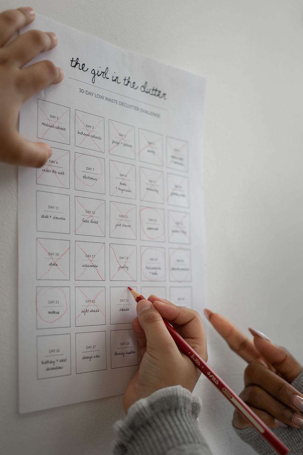 Hands mark a 30-day low-waste declutter challenge worksheet titled the girl in the clutter, with red pencil and crossed-out days.