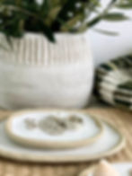 A small dish of jewellery lies on a woven placemat with a textured white pot containing a plant in the background.