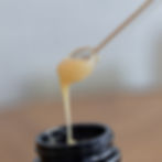 A wooden stick with thick, yellowish liquid being lifted from an open dark jar against a blurred background.