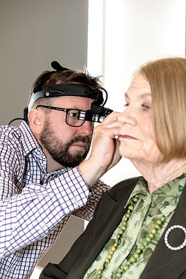 An optometrist examines an elderly woman's eye using a head-mounted device in a well-lit room.