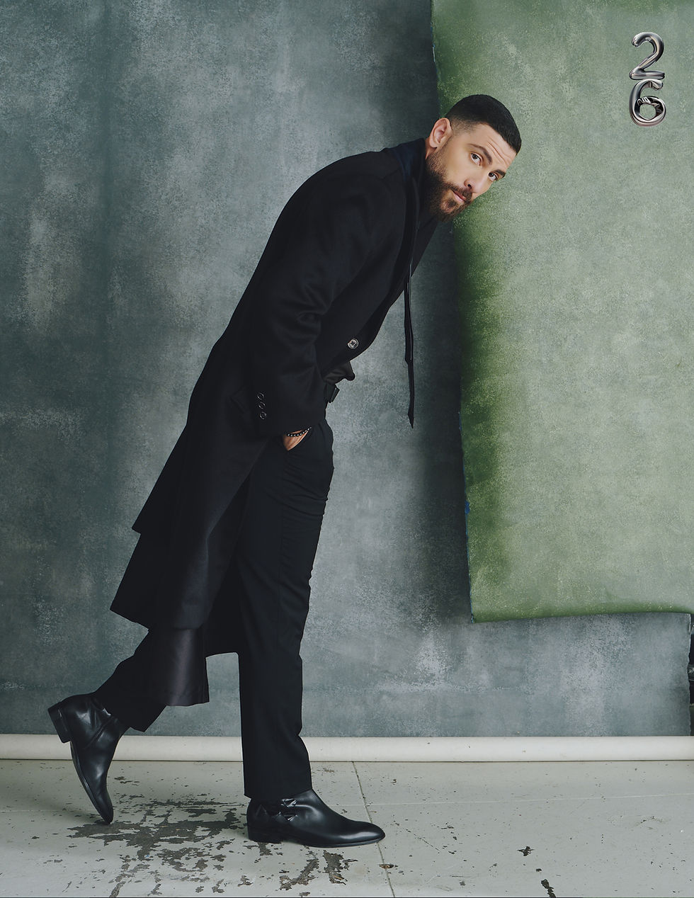 Zeeko Zaki in front of a weathered grey wall, wearing a long black duster coat and boots, captured by 26 Magazine
