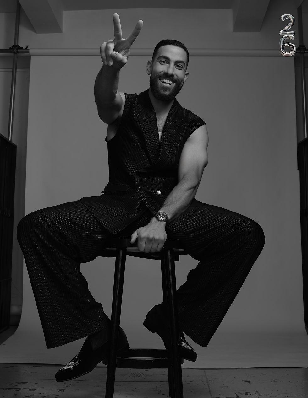 leading man Zeeko Zaki smiling brightly while holding up a peace sign, seated in a sleeveless vest, for 26 Magazine's cover story
